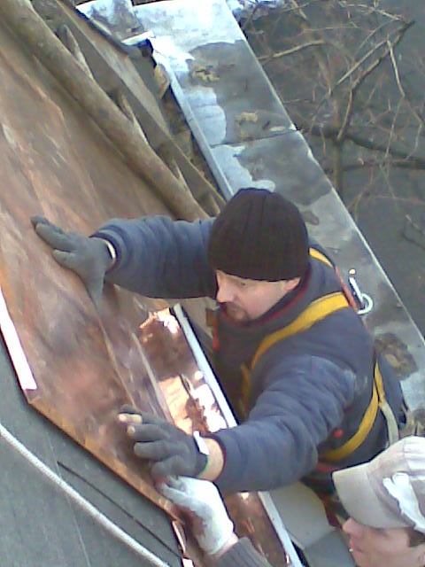 Dekarz w czapce i rękawicach roboczych montuje miedziane arkusze na dachu, zabezpieczony uprzężą, widok z dołu.